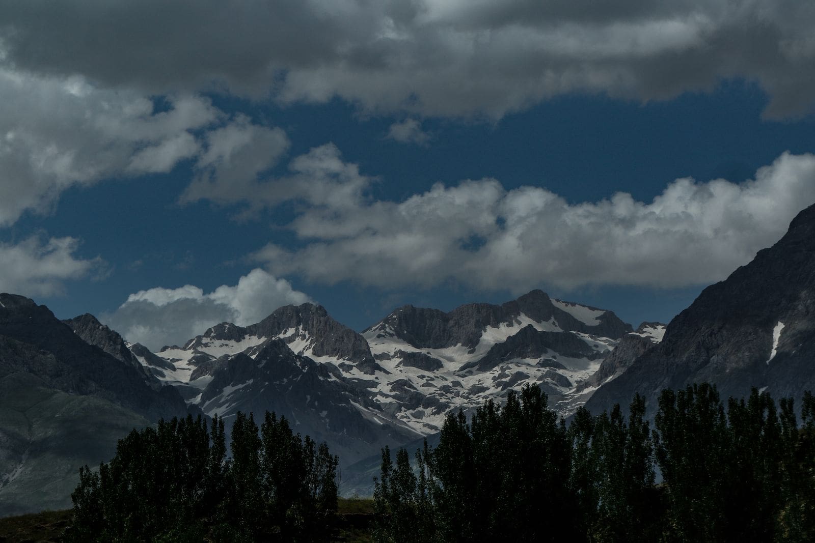 Snowcapped mountain peaks under dramatic clouds with a green tree line below, evoking Psalm 46:1 — God is our refuge and strength.