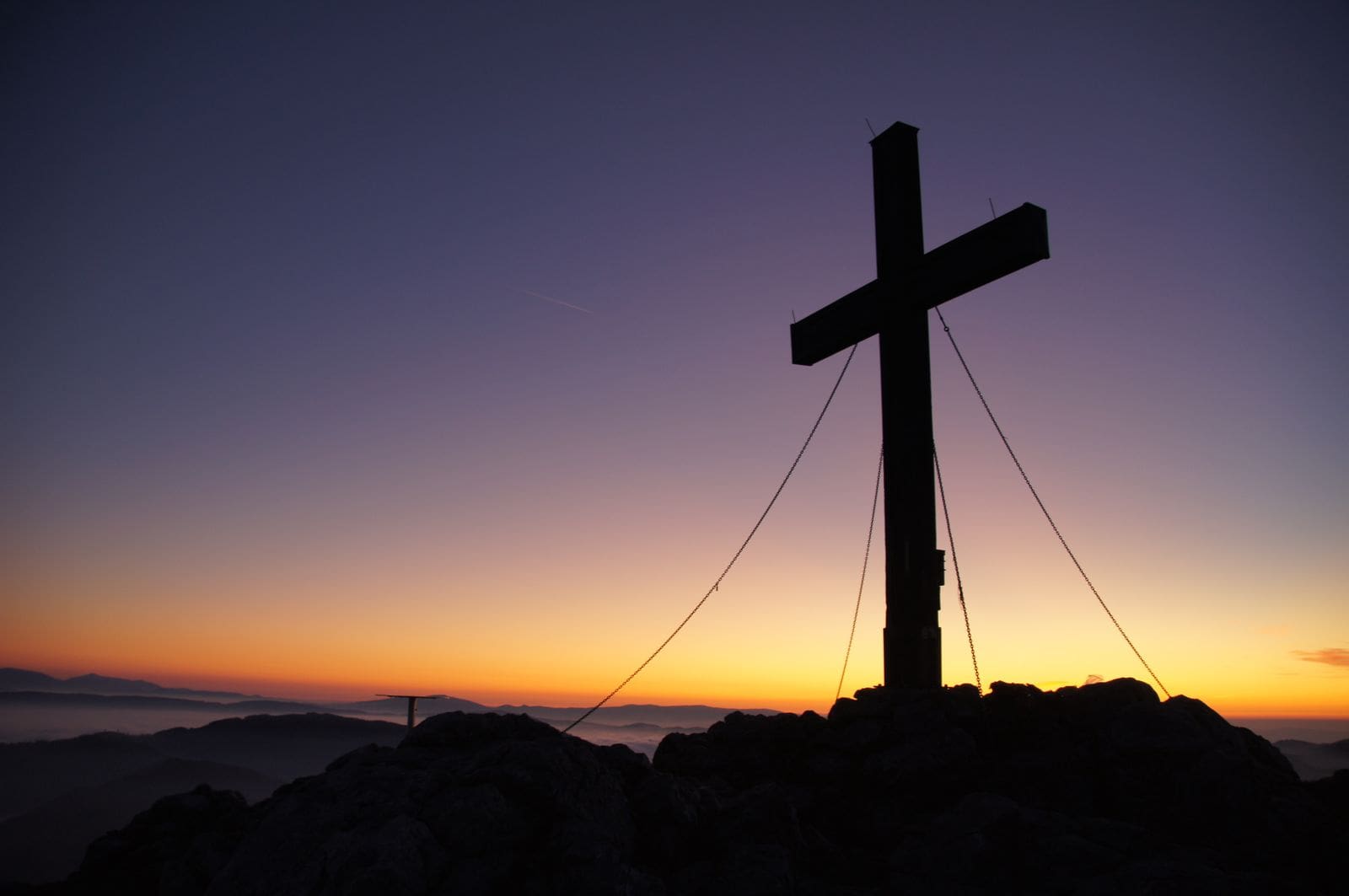 Wooden cross silhouetted against a dramatic golden sunset over mountain peaks — Jesus paid the debt of sin and rose so you would know it is true