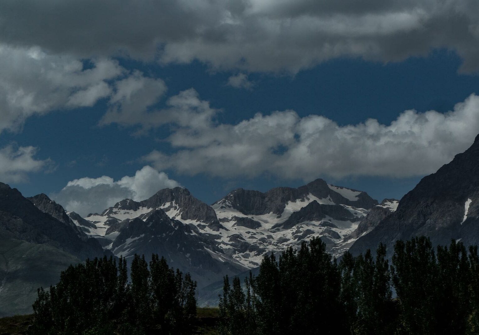 Snowcapped mountain peaks under dramatic clouds with a green tree line below, evoking Psalm 46:1 — God is our refuge and strength.