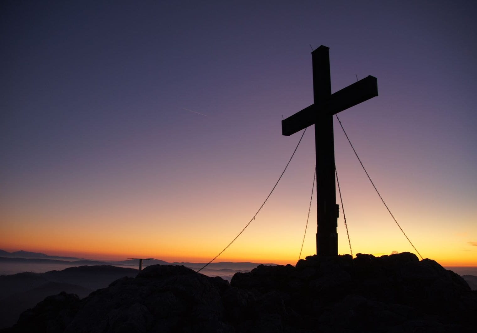 Wooden cross silhouetted against a dramatic golden sunset over mountain peaks — Jesus paid the debt of sin and rose so you would know it is true